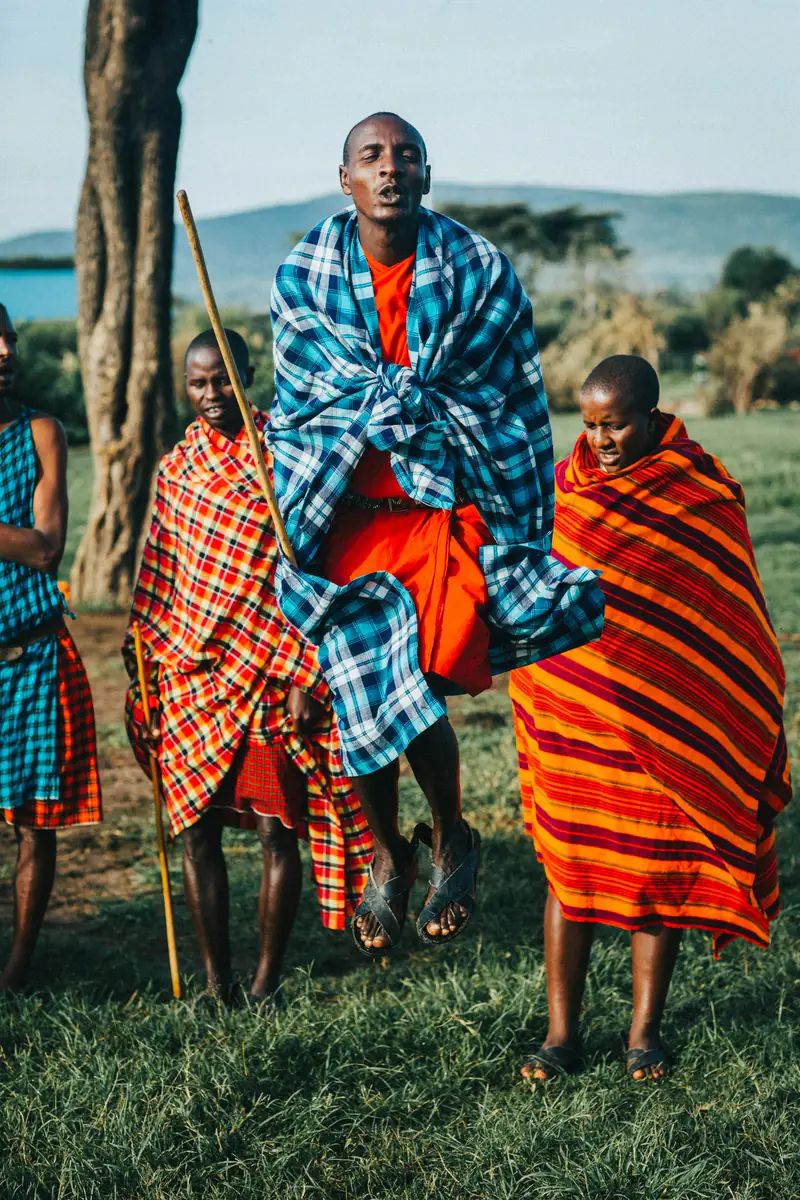 Maasai men in bright shukas, one mid-jump in an adumu dance, on open grass